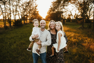 Mom and dad holding daughter and sun with golden sunset and trees behind them