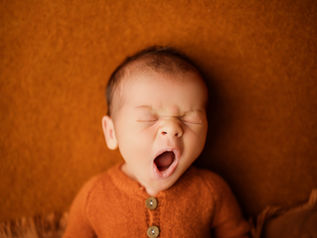 A newborn baby with dark brown hair yawning on a rust-colored fleece