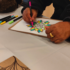 A person colouring a geometric mandala design on a clipboard, using a pink marker, with several coloured markers scattered on the table nearby.