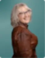 Portrait of a Jessica Malo, owner of Creekside Cabin Bookkeeping with shoulder-length wavy blonde-gray hair and glasses, wearing a brown leather jacket and posing against a teal background.