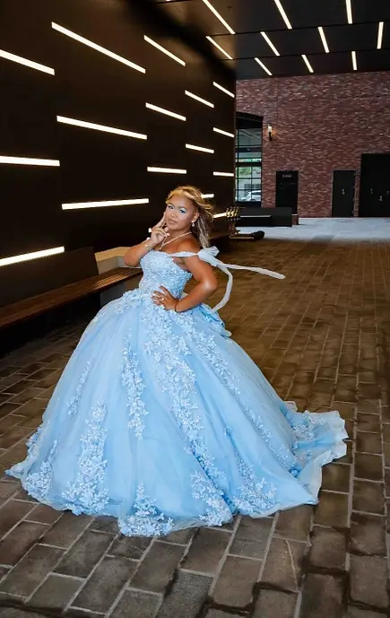 Sweet 16 portrait of a teen in a light blue floral ball gown standing in a modern hallway with dark walls, horizontal light strips, and brick flooring.