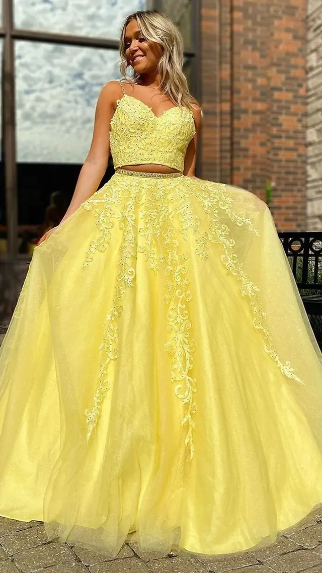 With blonde hair flowing, a Sweet 16 celebrant wears a lively yellow two-piece dress, posing against a brick wall with a window in the background. The bold color of the dress pops against the urban setting, adding a modern touch to her celebration look.