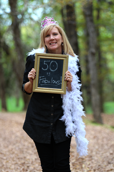 The 50th birthday celebrant is wearing a tiara, a black shirt and pants, and a white scarf. She holds a sign that says, '50 is Fabulous!' while smiling and posing for a photograph.