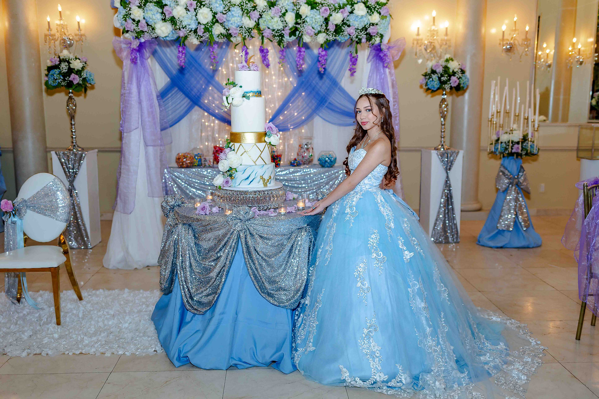 Sweet 16 birthday girl posing beside her three‑tier cake while wearing a stunning blue gown at her New Jersey celebration.