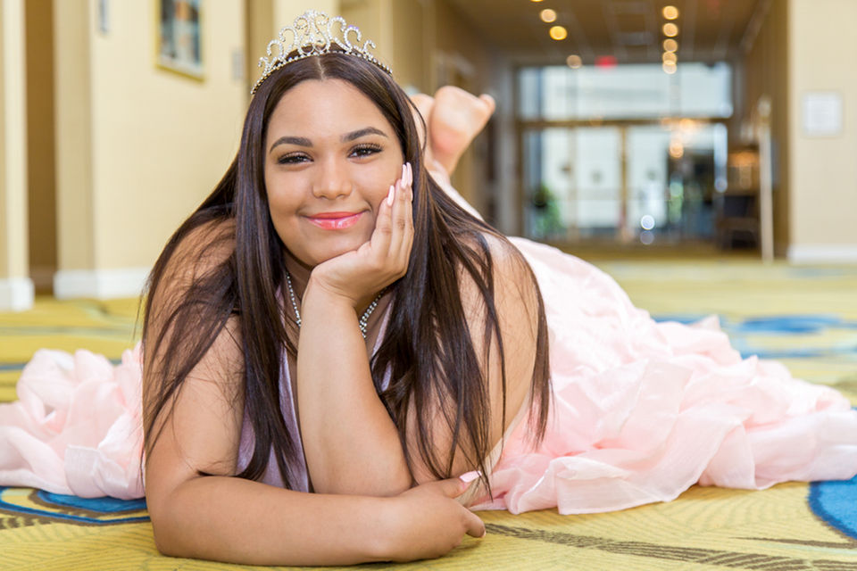 The Quinceañera birthday girl strikes a graceful pose while lying on the floor. Her lustrous black hair cascades, complementing her elegant peach dress.