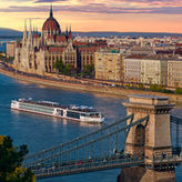 Vista aérea del barco fluvial Viking Eldir navegando por el río Danubio frente al Parlamento de Budapest al atardecer, con el Puente de las Cadenas en primer plano y el cielo teñido de tonos rosados y dorados.
