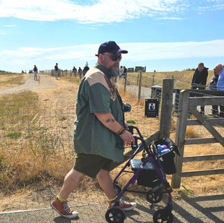 Bob using his rollator walking the site around stonehenge. This was taken during a heatwave so the grass is dry and yellowed. The sky behind him is blue. He wearing shorts and tshirt with an open shirt, cap and sunglasses.
