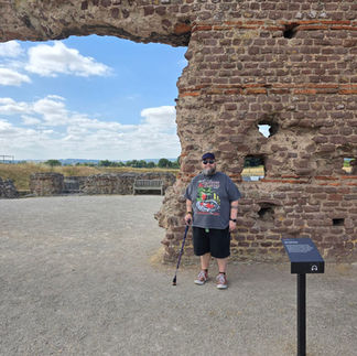 An image of Bob standing next to the archway. He is wearing a t-shirt, shorts and cap and using his walking stick.