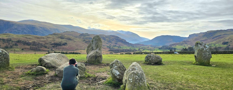 Our son, crouching to take photographs at Castlerigg Stone Circle with mountains in the background.
