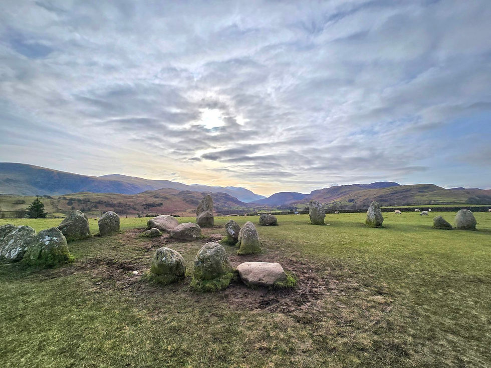 A view of Castlerigg stone circle/ The photo is taken from the centre showing the smaller inner circle. Behind the stones the hills and mountains of the lake district can be seen. The sky is cloudy and grey but pleasant. The ground is muddy.