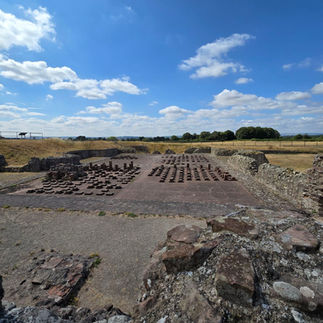 A view across Wroxeter Roman city showing the excavated ruins of the bath house that once stood here. The ruins are stone and low to the ground outlying the builds that were once here. The sky is blue with some wispy white clouds.