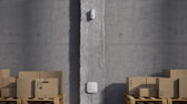 White Ajax Wireless MotionCam installed on grey concrete wall in warehouse, with pallets stacked with cardboard boxes in background.