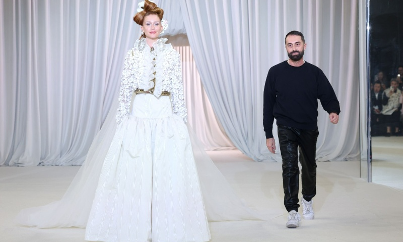 Uma modelo caminha por uma passarela vestindo um look branco de alta-costura, composto por uma saia volumosa, um casaco bordado e um véu longo. Seu cabelo está preso em um penteado elegante com um adorno dourado. Ao fundo, um estilista de barba e cabelos curtos, vestindo um suéter preto e calças brilhantes, caminha atrás dela. O cenário tem cortinas brancas drapeadas e um chão claro, criando uma atmosfera sofisticada e etérea.