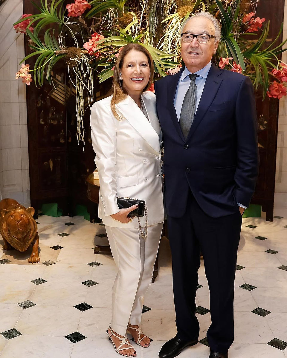 Donata Meirelles e Nizan Guanaes posando juntos em um ambiente sofisticado com decoração exuberante. Donata veste um elegante terno branco com sandálias de salto e segura uma clutch preta, enquanto Nizan usa um terno azul escuro com gravata cinza. O cenário ao fundo apresenta flores tropicais e elementos decorativos luxuosos, incluindo uma escultura de leão no chão. Ambos sorriem, transmitindo elegância e confiança.