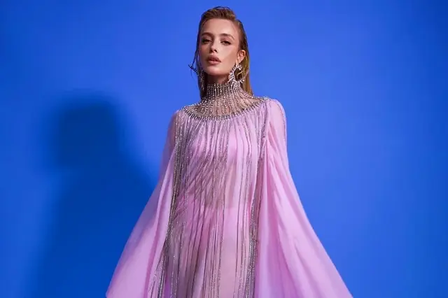 Maria Braz no Carnaval Gala da PatBO na NYFW – Maria Braz deslumbra em um vestido rosa fluido, com franjas de cristais e um colar de pedrarias que destaca seu pescoço, criando um efeito de realeza moderna contra o fundo azul.