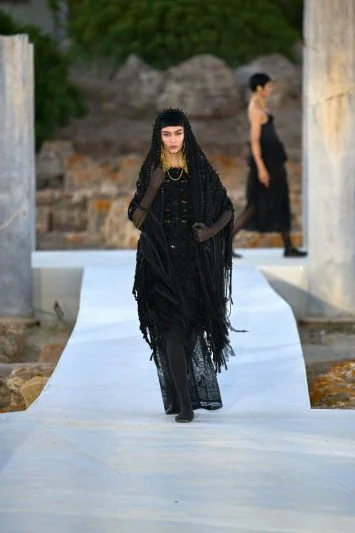 Modelo desfilando em uma passarela ao ar livre, vestindo um traje preto dramático com franjas e detalhes texturizados, combinado com um véu longo e acessórios dourados. O cenário de ruínas antigas ao fundo e outra modelo em destaque criam um contraste entre o contemporâneo e o histórico, adicionando intensidade à apresentação