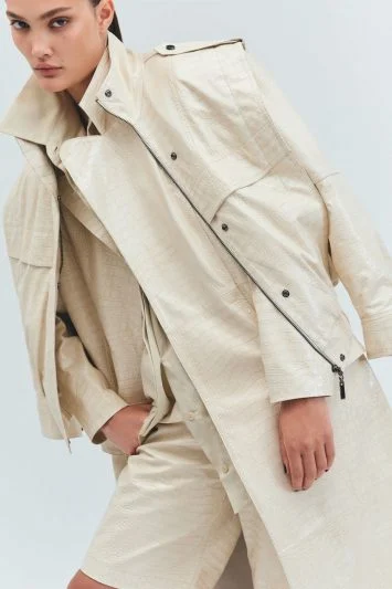 Modelo com olhar intenso veste um look monocromático em tom bege texturizado, composto por um trench coat oversized, blazer e bermuda estruturada. A pose inclinada e o zíper do casaco parcialmente aberto adicionam um toque de atitude ao visual minimalista e sofisticado.