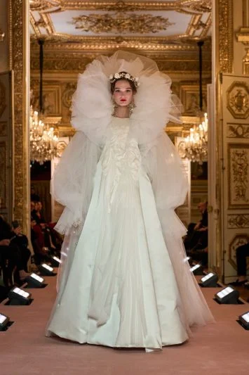 Modelo vestindo um exuberante vestido branco de alta-costura com camadas de tule e mangas volumosas, desfilando em um salão ricamente decorado, evocando uma noiva etérea e majestosa.
