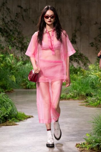 Uma modelo caminha pela passarela durante um desfile da Gucci em Londres, usando um conjunto rosa transparente composto por camisa e saia midi. O look é completado com um top rosa por baixo, bolsa vermelha pequena, sapatos brancos de plataforma, óculos de sol avermelhados e colar de pérolas. A passarela é decorada com vegetação exuberante e o cenário tem um aspecto natural e moderno.