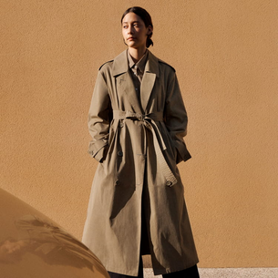 Modelo usando um trench coat verde militar de algodão com amarração na cintura e detalhes de botões duplos, em um cenário de parede texturizada em tom terroso.