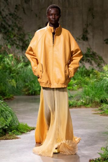 Uma modelo desfila com um look da Gucci em Londres, composto por uma jaqueta oversized em tom amarelo-mostarda e uma saia longa plissada, fluida e transparente no mesmo tom, que se arrasta pelo chão. O cenário da passarela é coberto de vegetação verde exuberante, criando um contraste natural com o visual monocromático da modelo.