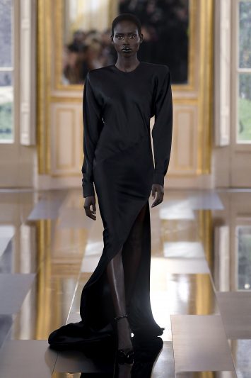 Uma modelo desfila com um elegante vestido preto longo de manga comprida, com uma fenda alta na perna e uma cauda fluida que se arrasta pelo chão. A silhueta do vestido é minimalista e sofisticada, destacando-se pela simplicidade do design. A modelo desfila com expressão séria, maquiagem escura nos lábios e sapatos pretos de salto alto, caminhando pela passarela espelhada em um cenário luxuoso com molduras douradas durante a Paris Fashion Week