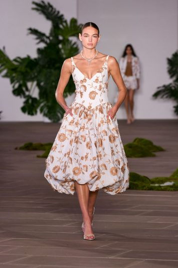 Modelo desfilando na passarela da coleção Ethereal da PatBo, usando um vestido midi branco com estampa floral em tons de dourado e saia volumosa estilo balonê. O look é complementado por um colar delicado, e o cenário ao fundo apresenta vegetação natural, criando uma atmosfera elegante e romântica.