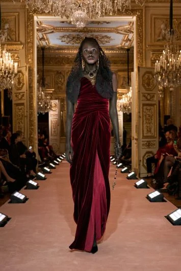 Modelo vestindo um vestido de veludo vinho drapeado, com acessórios de inspiração medieval, caminhando com imponência pelo ambiente luxuoso da passarela.