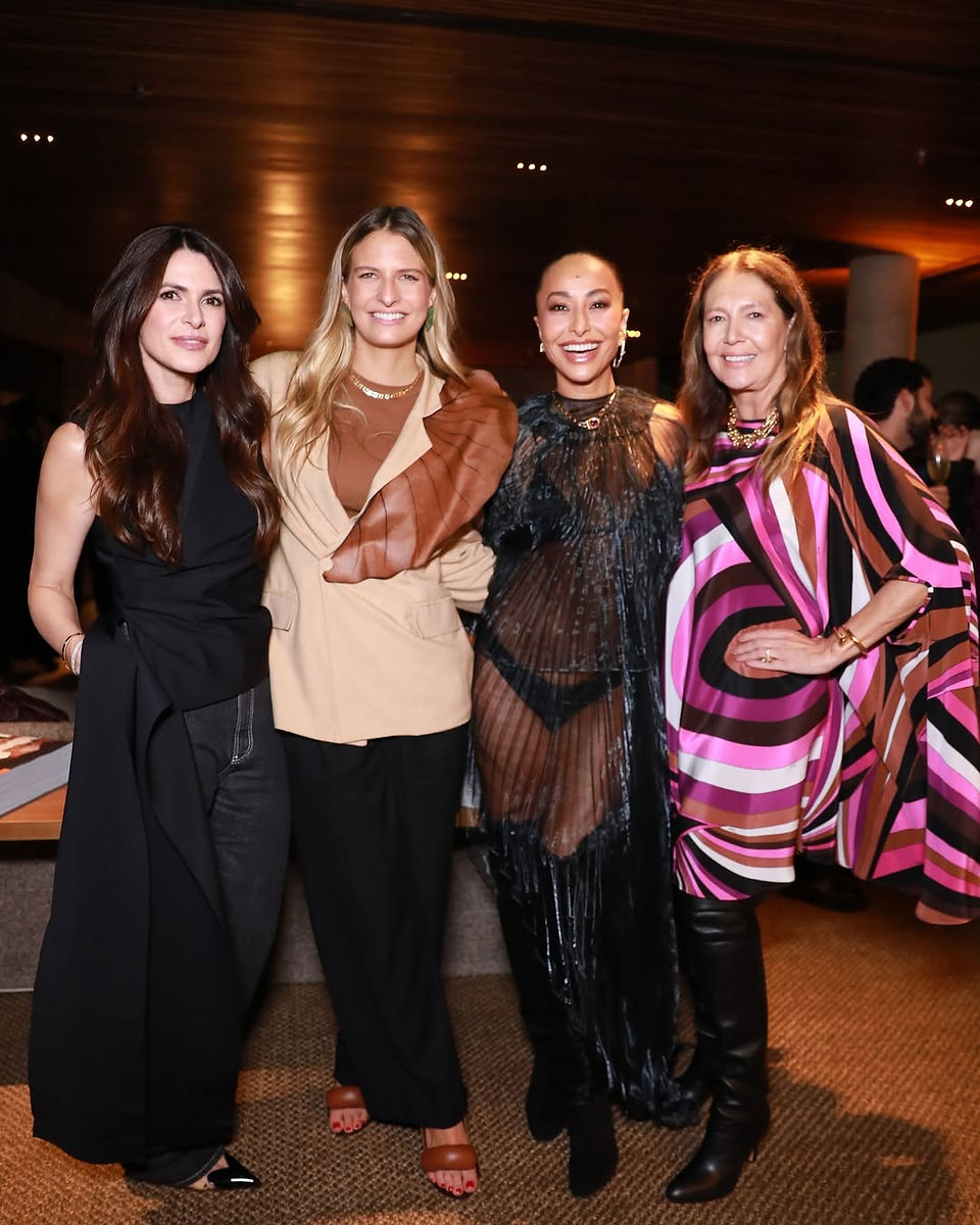 Quatro mulheres posam juntas em um evento elegante, com iluminação quente ao fundo. Da esquerda para a direita: uma mulher vestindo preto com cabelo solto, outra com blazer bege e detalhe marrom, uma terceira usando um vestido preto translúcido e colar dourado, e Donata Meirelles com uma blusa de estampas geométricas em tons de rosa, marrom e preto, acompanhada de acessórios dourados. Todas sorriem, transmitindo sofisticação e carisma.