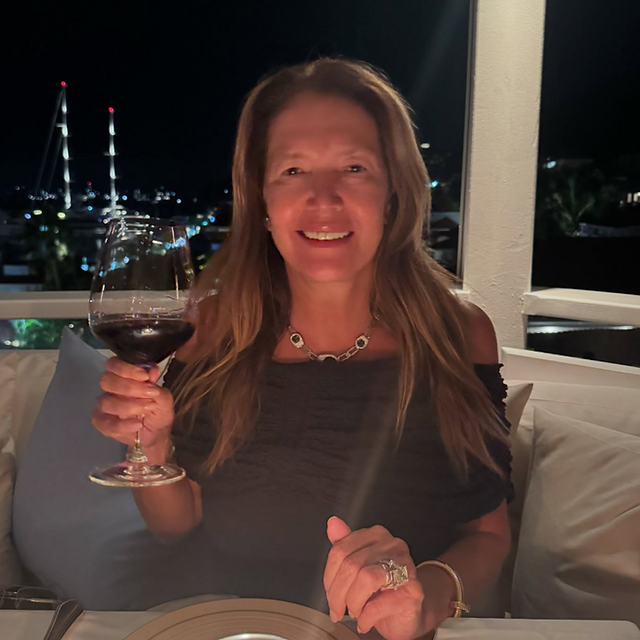 Donata Meirelles brinda com uma taça de vinho tinto em um jantar ao ar livre durante a noite. Ela veste um elegante vestido preto ombro a ombro e um colar refinado, com um cenário iluminado e um barco ao fundo.