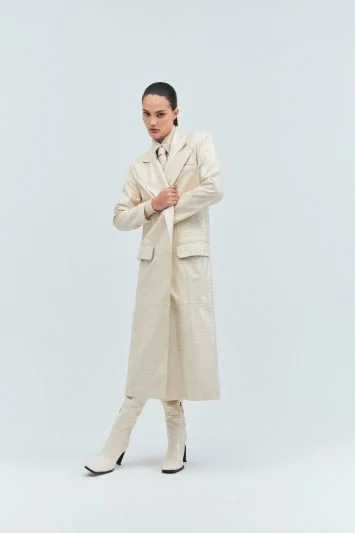 Modelo com postura elegante veste um longo trench coat bege texturizado, combinado com calça reta e botas de bico fino no mesmo tom. A gravata fina adiciona sofisticação ao look monocromático, que reflete uma estética moderna, minimalista e sofisticada. O fundo claro destaca a imponência da silhueta estruturada.