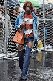 Modelo desfilando com uma Telfar Bag marrom em um look casual com jaqueta jeans e botas de cano alto, em um ambiente urbano e chuvoso.