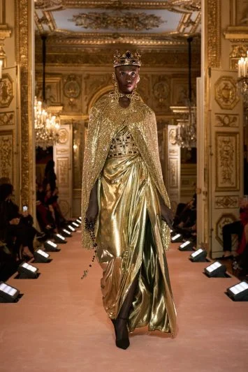 Modelo deslumbrante em um vestido dourado metálico, acompanhado de uma capa brilhante e uma coroa, desfilando com imponência em uma passarela rodeada de lustres clássicos e paredes ornamentadas.