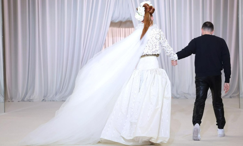 Vista de costas, uma modelo caminha segurando a mão de um estilista, que também está de costas. Ela veste um look branco de alta-costura com uma saia volumosa e um véu longo esvoaçante, que se estende atrás dela. O casaco bordado e o cinto metálico acrescentam detalhes sofisticados ao visual. O estilista usa um suéter preto e calças brilhantes, combinando com o cenário minimalista de cortinas brancas drapeadas, que criam uma atmosfera etérea e elegante.