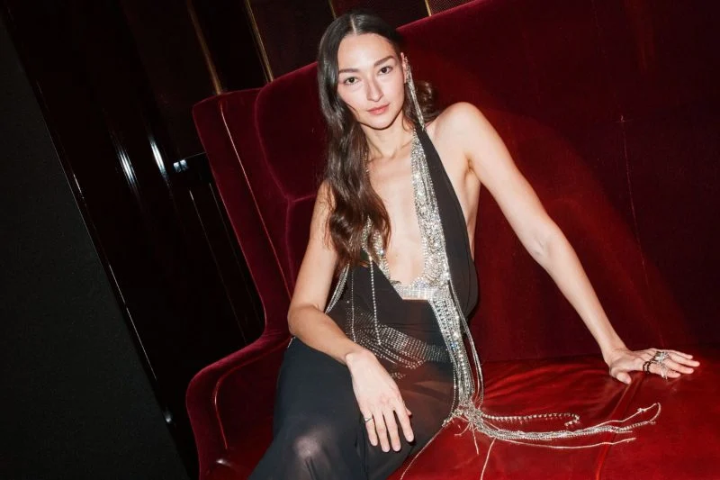 Modelo Bruna Tenório posa em um sofá de veludo vermelho usando um vestido preto profundo com detalhes prateados e transparência, trazendo um ar sofisticado e sensual ao look.