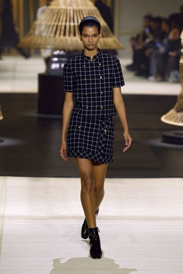 Modelo desfilando na passarela da Dior com conjunto preto de estampa quadriculada branca, composto por blazer de abotoamento duplo e saia curta, combinado com botas pretas e cenário com estruturas de junco iluminada