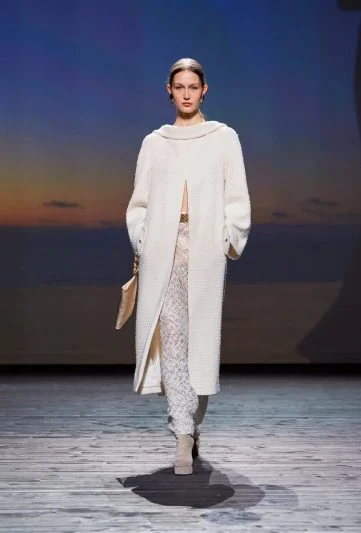 Modelo no desfile Chanel FW 2024/25, vestindo um elegante casaco branco oversized combinado com calça rendada branca, botas bege e bolsa de mão, desfilando em uma passarela com cenário de pôr do sol no mar.