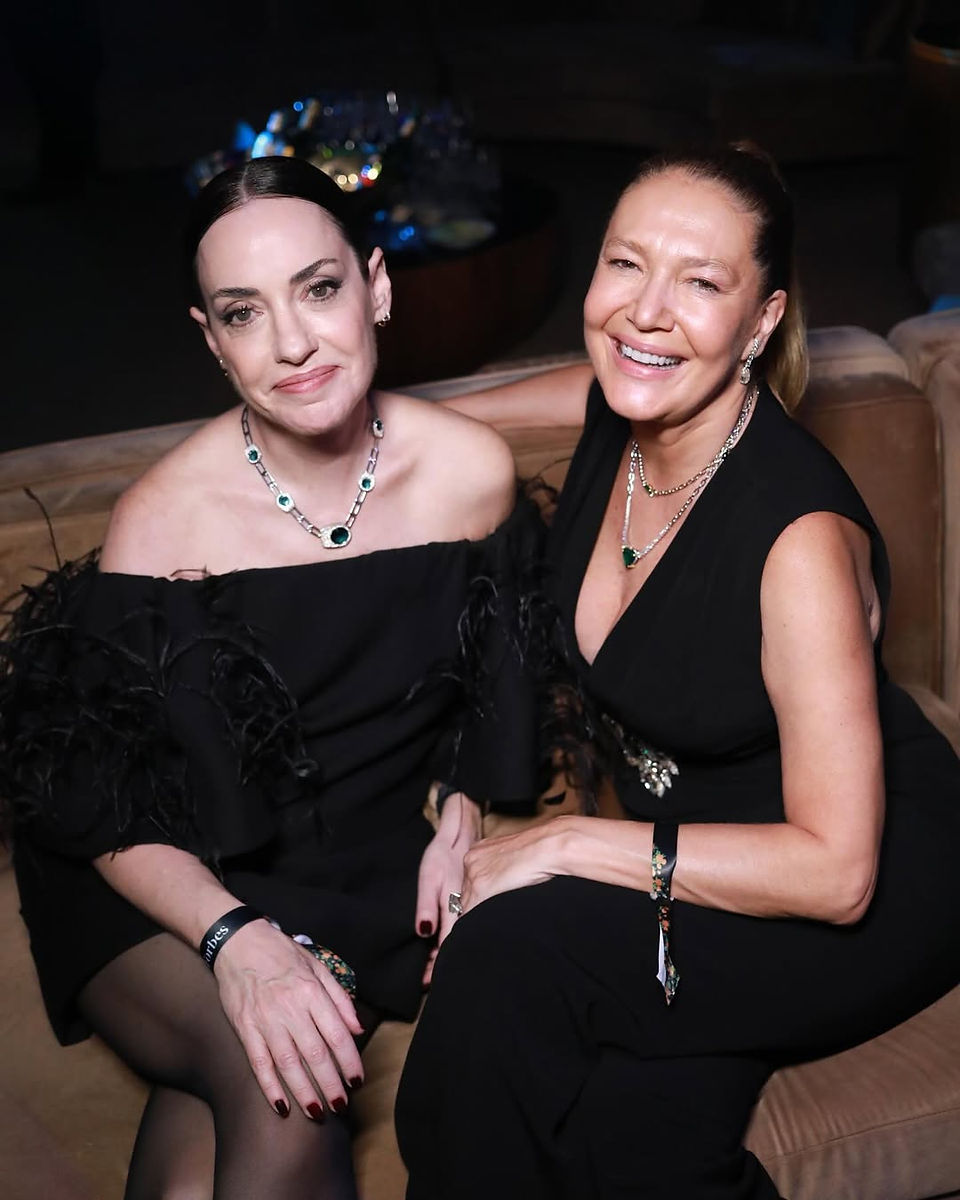 Donata Meirelles ao lado de uma amiga em um evento elegante. Ambas usam vestidos pretos sofisticados e joias marcantes, com sorrisos descontraídos em um ambiente acolhedor