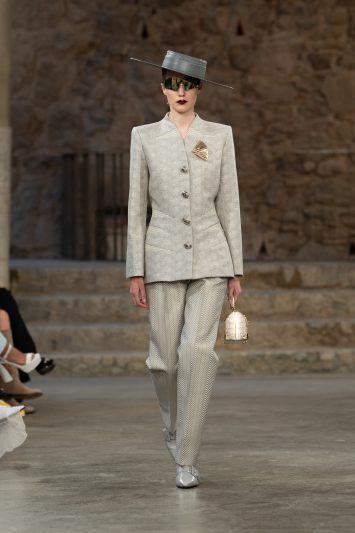 Modelo caminhando em uma passarela vestindo um conjunto elegante de alfaiataria cinza claro com padrão discreto, composto por blazer de corte reto com botões frontais e calça de pernas retas. O visual é complementado por um chapéu geométrico cinza de aba fina, óculos de sol espelhados e um broche dourado. A modelo segura uma pequena bolsa estruturada dourada em uma das mãos. O cenário ao fundo exibe paredes de pedra e degraus, conferindo uma atmosfera rústica e refinada
