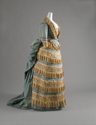 Vestido de noite do final do século XIX criado por Charles Frederick Worth, com saia drapeada verde e detalhes em franjas douradas, refletindo a estética luxuosa da Belle Époque.