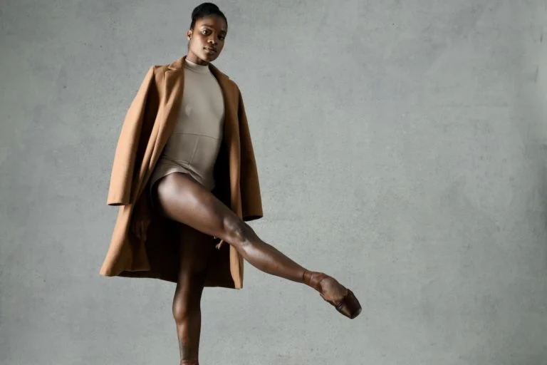 A bailarina Ingrid Silva posa com elegância contra um fundo neutro, vestindo um collant nude, casaco camel e sapatilhas de ponta. Sua postura forte e confiante reflete sua trajetória de disciplina e superação no balé.