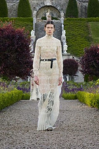 A imagem apresenta uma modelo desfilando em um jardim formal, vestindo um vestido longo de renda branca com gola alta e mangas compridas, transmitindo uma estética romântica e etérea. A cintura é marcada por um cinto preto que adiciona contraste e modernidade ao visual delicado. O cenário ao fundo, com escadarias e vegetação simétrica, destaca o equilíbrio entre o clássico e o contemporâneo.