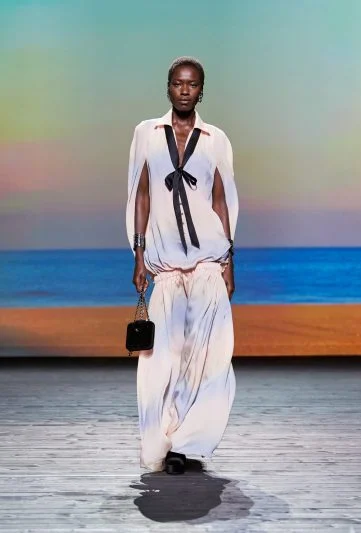 Modelo desfilando na coleção Chanel FW 2024/25, usando um vestido longo fluido em tons claros com laço preto na gola, acessórios metálicos e bolsa de mão, sobre uma passarela com cenário de praia ao pôr do sol