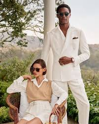 Casal vestindo looks impecáveis inspirados na estética "Old Money". O homem usa um terno branco de corte clássico, enquanto a mulher aposta em um colete bege sobre camisa branca e saia clara. Ambos usam óculos escuros sofisticados, reforçando a elegância atemporal do estilo.