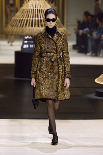 Modelo desfilando na passarela da Dior com casaco de estampa de onça ajustado na cintura com cinto, meia-calça preta, sapatos clássicos e óculos de sol escuros, em cenário com estruturas de junco iluminadas