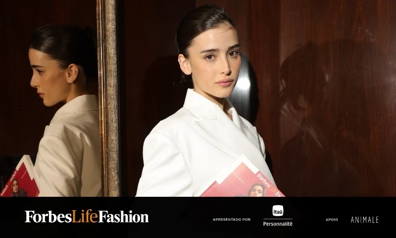 Livia Nunes posa para a foto. Ela usa um terninho branco e carrega revistas da forbes, de capa vermelha, nas mãos.