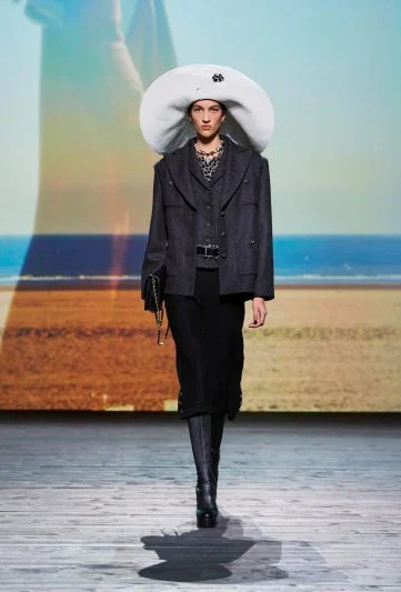 Modelo no desfile Chanel FW 2024/25, vestindo um conjunto preto com blazer estruturado, saia midi e botas altas, complementado por um chapéu branco oversized e colar statement, desfilando em uma passarela com cenário de praia ao pôr do sol