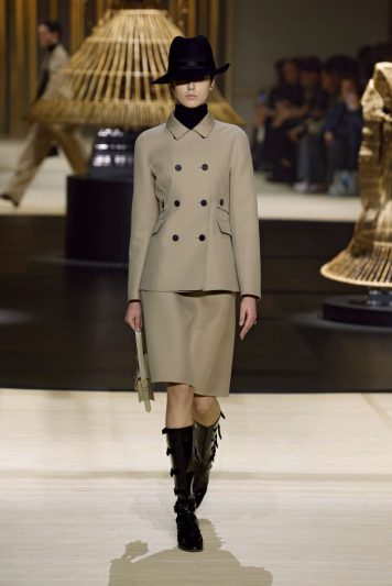 Modelo desfilando na passarela da Dior com look composto por casaco bege estruturado de abotoamento duplo, saia midi combinando, botas de cano alto pretas com detalhes e chapéu preto, em cenário decorado com estruturas de junco iluminadas.