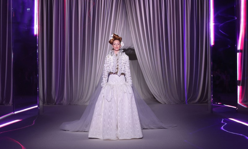 Uma modelo está parada no centro da passarela, usando um look branco de alta-costura. O conjunto inclui uma saia volumosa, um casaco bordado e um longo véu que se estende pelo chão. Seu cabelo está preso em um penteado elaborado, adornado com um acessório dourado. O cenário é sofisticado, com cortinas brancas drapeadas ao fundo e luzes neon roxas refletindo nas laterais, criando um contraste moderno e futurista com o visual clássico da modelo.