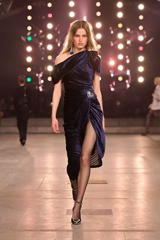  Modelo usando um vestido de veludo azul marinho com um ombro só, com detalhe de broche e fenda lateral, combinado com sandálias de tiras.